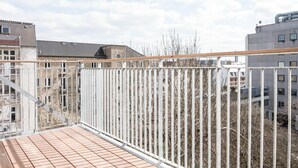 Un balcone con ringhiera in metallo, pavimento piastrellato e vista su edifici e alberi spogli sullo sfondo.