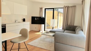 Una sala de estar moderna con un sofá gris, un televisor de pantalla plana, una mesa de comedor con una silla y una zona de cocina con armarios blancos y un televisor negro.