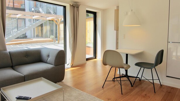 Una sala moderna con un sofá, una mesa de centro y dos sillones. Hay un ventanal con vistas a una escalera y un edificio.