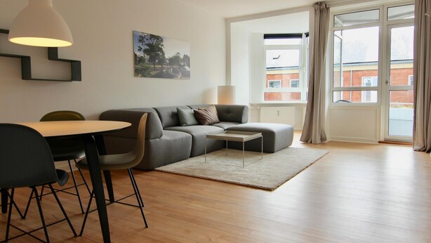 Ett modernt vardagsrum med en soffa, ett matbord och stolar, en matta och ett stort fönster med gardiner.
