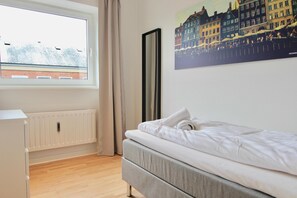 A bedroom with a bed, a radiator, a window, and a cityscape painting on the wall.