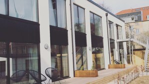 Un bâtiment moderne doté de grandes fenêtres, d’une terrasse en bois et d’un espace salon extérieur.