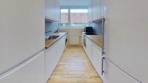 A narrow kitchen with white cabinets, a wooden countertop, a washing machine, and a window with a view of buildings. - 3-bed in Frederiksberg w Balcony - 210 (Frederiksberg)