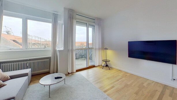A modern living room with a sofa, a coffee table, a TV, and a balcony with a view of buildings. - 3-bed in Frederiksberg w Balcony - 210 (Frederiksberg)