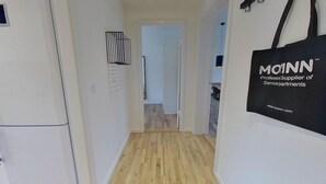 A hallway with wooden flooring, a white wall, and a door leading to a room. A black bag with the text "MOINN Professional Supplier of Servicial/partments" hangs on the wall. - 3-bed in Frederiksberg w Balcony - 210 (Frederiksberg)