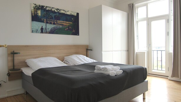 A bedroom with a large bed, a nightstand, a painting of a riverside scene, a wardrobe, and a large window with curtains. - Beautiful 2-bed in Frederiksberg C W Balcony (Frederiksberg)
