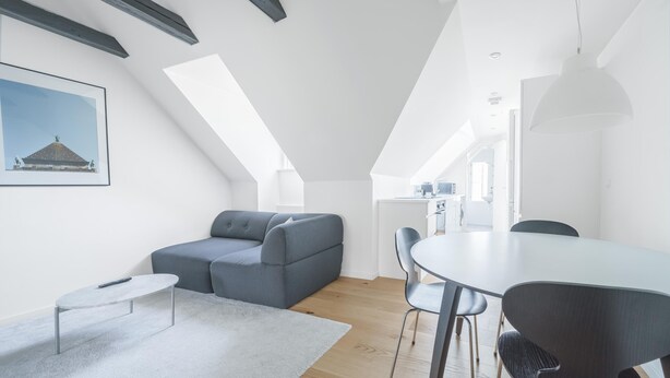 A modern loft apartment with a loft bed, a gray sofa, a dining table with chairs, and a kitchen area in the background. - Beautiful 1-bed in Peaceful Surroundings (Aarhus)