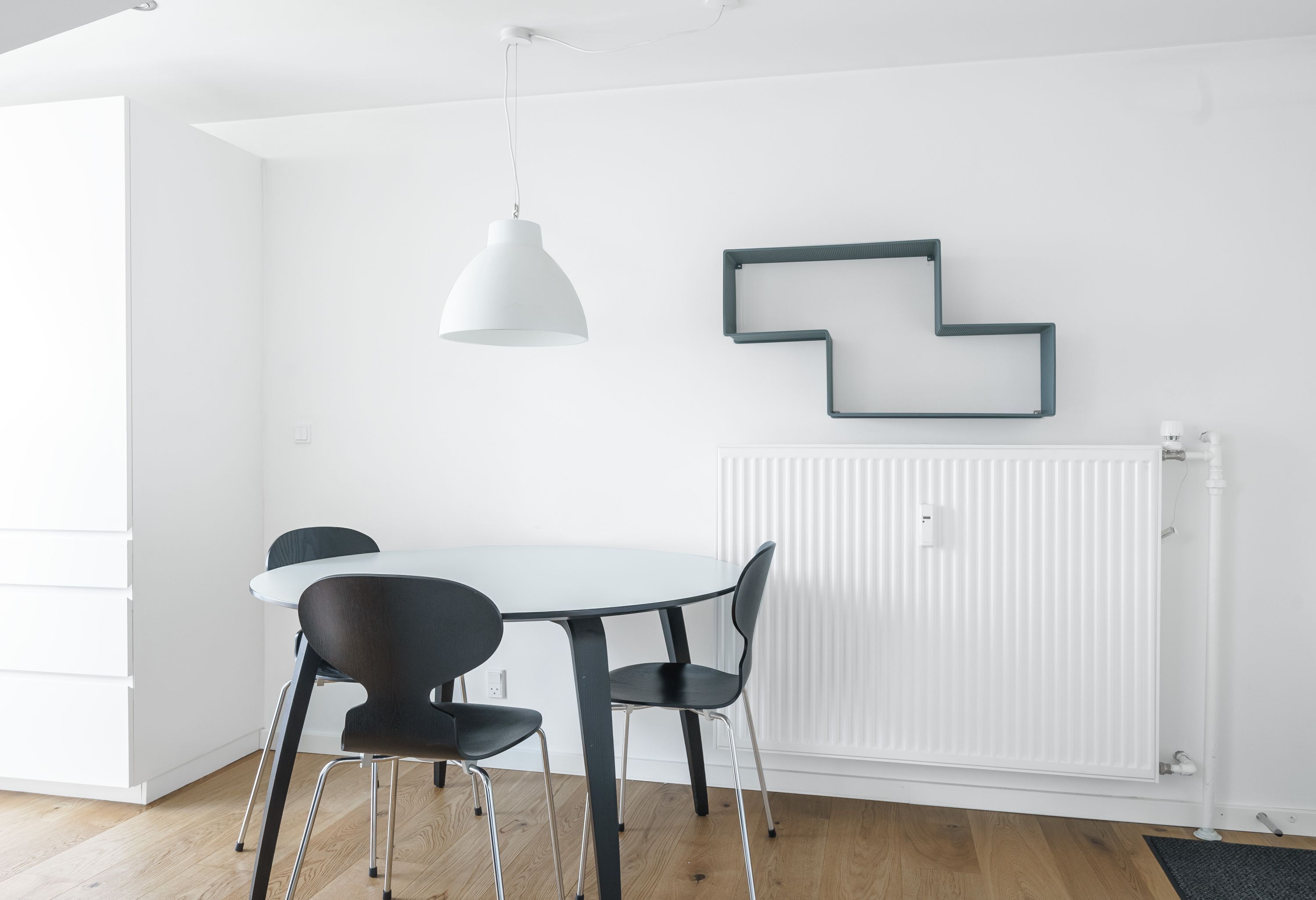 Un espace de salle à manger moderne avec une table blanche, des chaises noires, un luminaire suspendu blanc et un élément décoratif géométrique sur le mur.