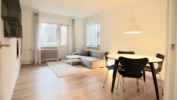 Ein modernes Wohnzimmer mit Sofa, Esstisch und Stühlen, großem Fenster mit Vorhängen sowie einer Pendelleuchte.