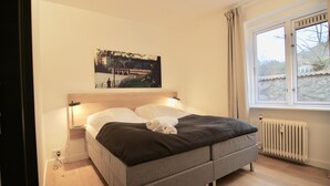 Ein Schlafzimmer mit einem Bett, einem Nachttisch, einem Wandbild, einem Fenster und einer Heizung.