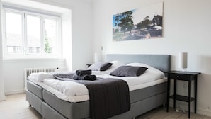 Una camera da letto moderna con un letto grigio, biancheria bianca, un comodino nero e una finestra con vista su una casa e degli alberi.