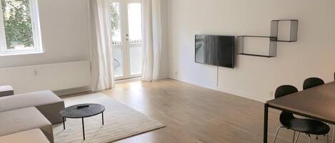 A spacious living area with a sofa, a coffee table, a TV mounted on the wall, and a dining table with chairs.