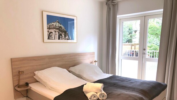 Ein Bett mit Holz Kopfteil, weißen Kissen und einer dunklen Decke. Darüber an der Wand hängt ein gerahmtes Bild.