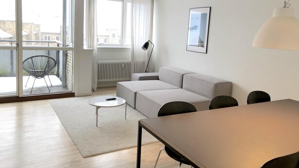A modern living room with a sofa, a dining table, and a chair. There is a large window with a view of buildings outside. - Nice 2-bed in Frederiksberg (Frederiksberg)