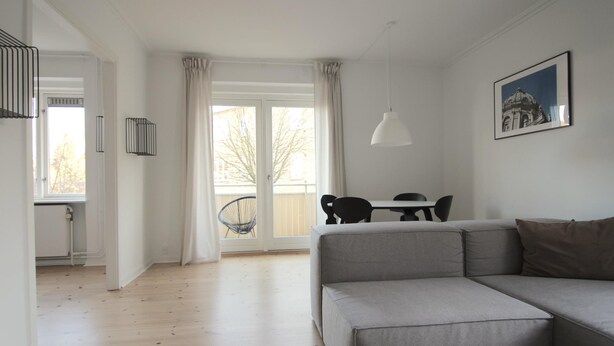 A modern living room with a grey sofa, a dining table with chairs, a large window with curtains, and a hanging pendant light. - Great 2-bed w Balcony in Charlottenlund (Charlottenlund)