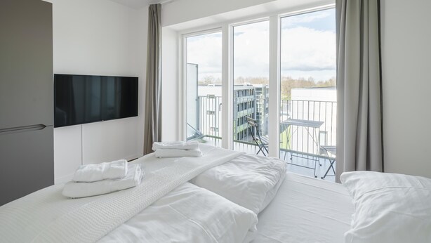 A hotel room with a bed, a flat-screen TV mounted on the wall, and a balcony with a view of the city.
