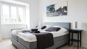Een moderne slaapkamer met een grijs bed, witte beddengoed, een zwart nachtkastje en een raam met uitzicht op een huis en bomen.
