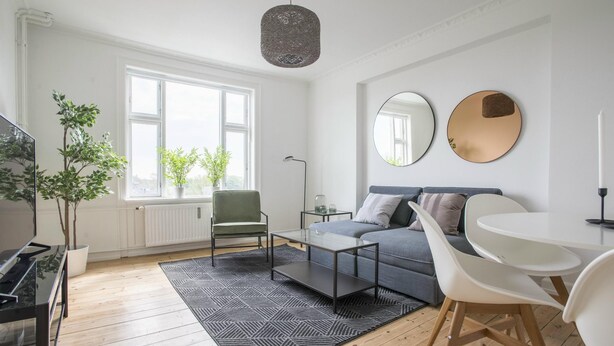 Una sala de estar moderna con sofá, mesa de centro y sillas. Se ve un ventanal con plantas, un radiador y una lámpara colgante.