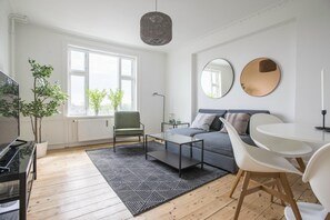 Moderni olohuone, jossa on sohva, sohvapöytä ja tuoleja. Siinä on suuri ikkuna, jonka edessä on kasveja, sekä patteri ja riippuvalaisin.