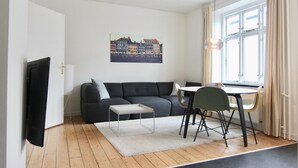 A modern living room with a grey sofa, a small coffee table, a dining table with chairs, a cityscape painting on the wall, and a window with curtains. - 2-bed Apartment in one of Rhus Best Locations (Aarhus)