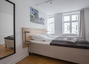 Une chambre à coucher moderne avec une tête de lit en bois, une grande fenêtre avec des rideaux, une table de chevet avec une lampe et un lit bien fait, agrémenté de serviettes pliées.