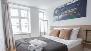 A neatly made bed with a grey and white checkered blanket, two brown pillows, and rolled white towels. A large window with white curtains allows natural light into the room. - Beautiful 2-bed in top Copenhagen Location (Copenhagen)