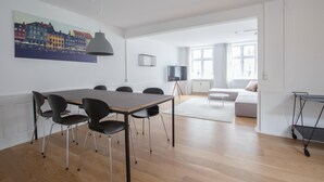 A modern living room with a dining area, featuring a wooden dining table, black chairs, a grey pendant light, a flat-screen TV, and a large window with curtains. - Beautiful 2-bed in top Copenhagen Location (Copenhagen)