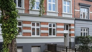 Un edificio residencial de dos pisos con fachada de ladrillo, ventanas con marco blanco y un pequeño patio delantero con cerramiento metálico.