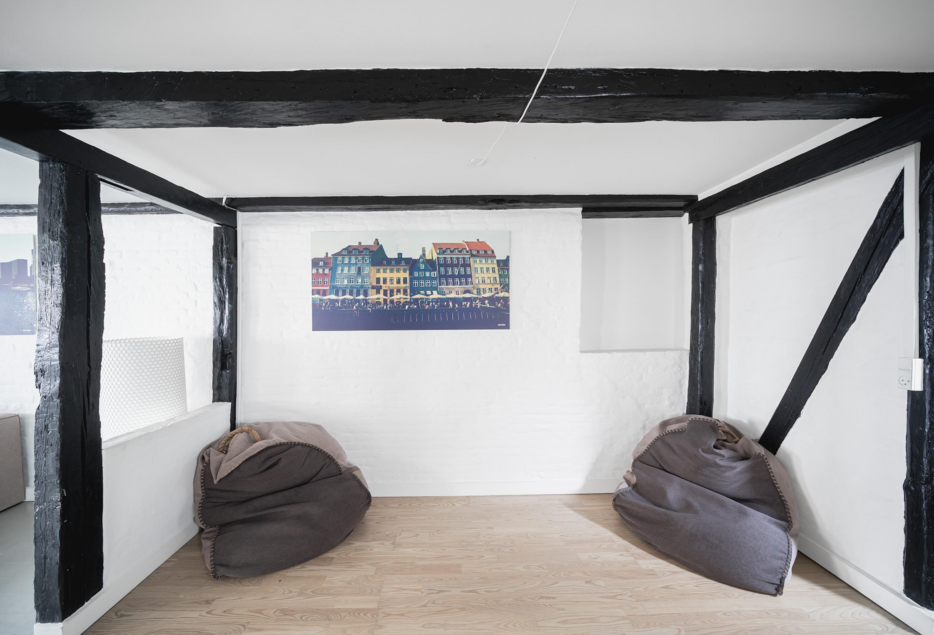 Ein Zimmer mit freiliegenden schwarzen Balken, einer weißen Wand und einem Holzboden. Zwei Sitzsäcke und ein gerahmtes Bild von einem bunten Gebäude sind vorhanden.
