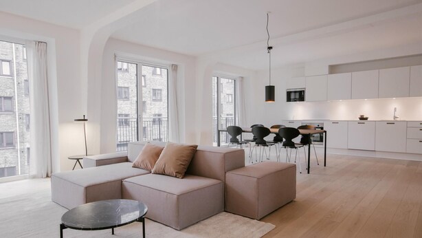 A modern living room with a sectional sofa, a round coffee table, and a dining area with a black chair and a small table. - Stunning 2-bed w Large Balcony (Copenhagen)