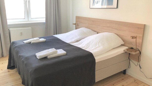 A neatly made bed with a grey bedspread and white pillows, a bedside table with a lamp, and a window with curtains. - Great Refurbished 2-bed in Amager Island (Copenhagen)