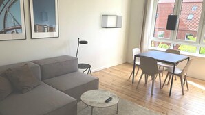 A modern living room with a grey sofa, a small round table, and a dining area with chairs. There are framed pictures on the wall and a window with a view of buildings. - Great Refurbished 2-bed in Amager Island (Copenhagen)
