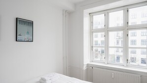 Una habitación de hotel con un ventanal grande, una cama con ropa de cama blanca, un radiador e un cuadro en la pared.