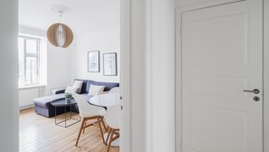Una sala de estar moderna con un sofá, una mesa redonda y sillas. Hay una puerta a la derecha y una ventana a la izquierda.