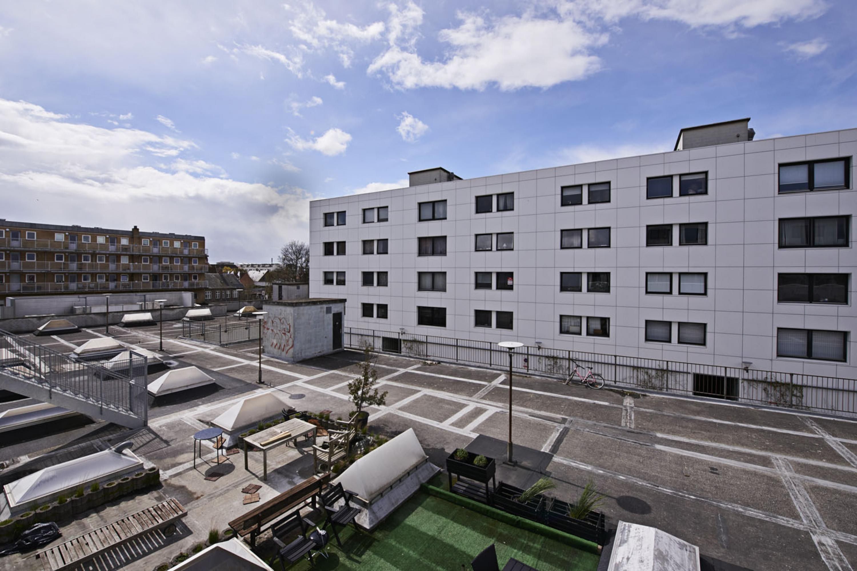 Een modern gebouw met een terras op het dak, buitenmeubels en een fiets nabij de ingang.