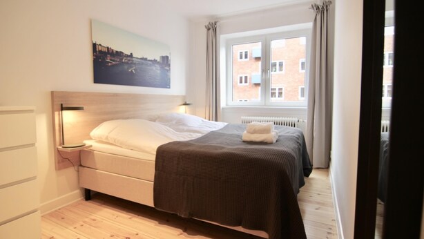 Ein Schlafzimmer mit einem Bett, einem Nachttisch, einem Fenster mit Vorhängen, einer Heizung und einem Stadtbild an der Wand.