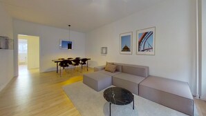 Ein modernes Wohnzimmer mit einem grauen Sofa, einem Esstisch mit Stühlen und einem schwarzen Couchtisch. An der Wand befinden sich zwei gerahmte Bilder.