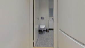 Ein enger Flur, der zu einem Badezimmer mit Toilette, Mülleimer und einem kleinen Waschbecken führt.