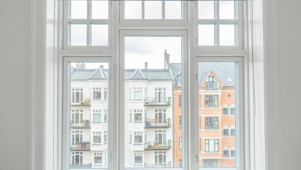 Une fenêtre à trois vitres, offrant une vue sur des immeubles à plusieurs étages dotés de balcons et d’une façade en briques.