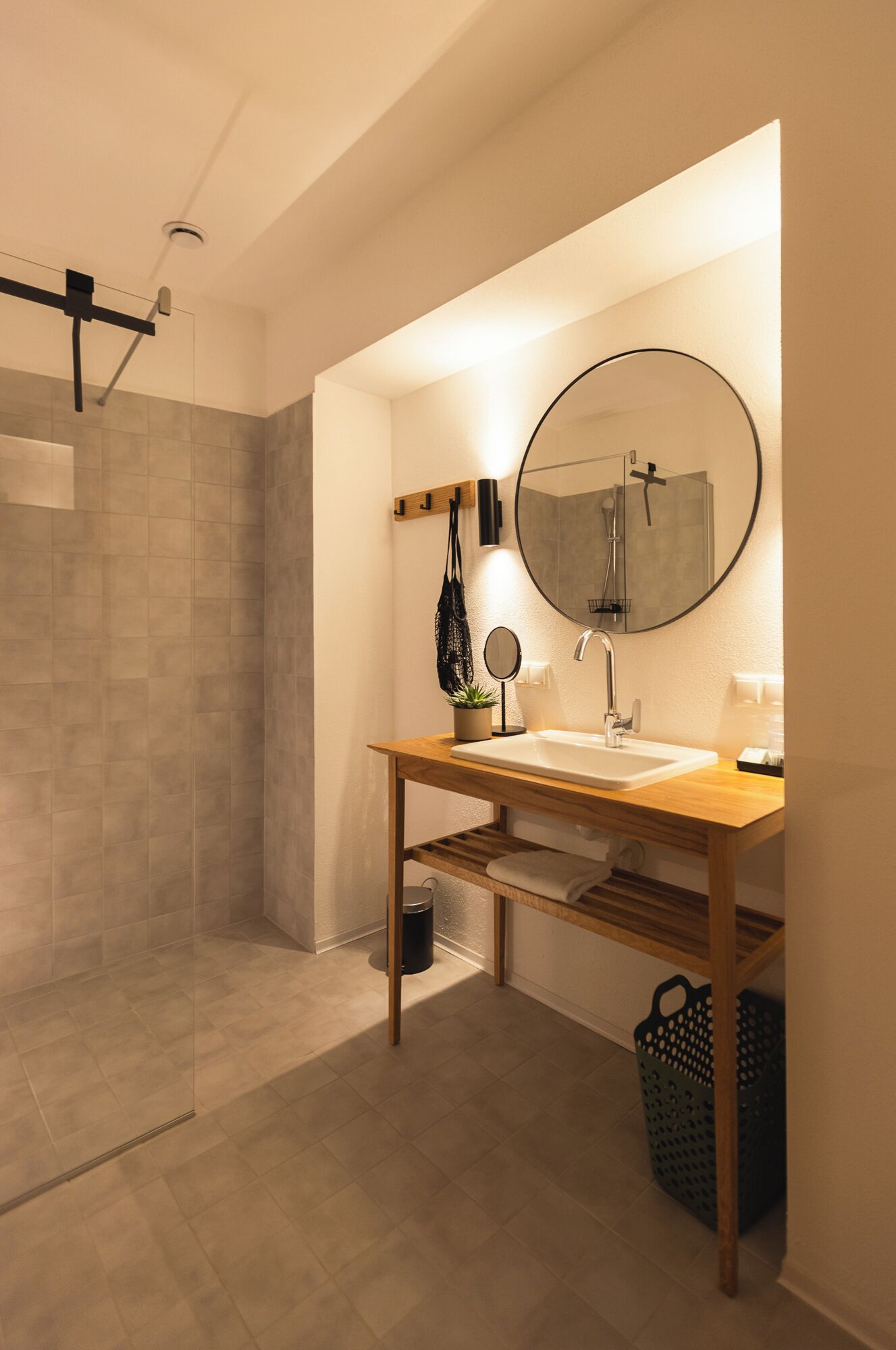 A modern bathroom with a wooden vanity, a round mirror, a shower, and a towel rack.