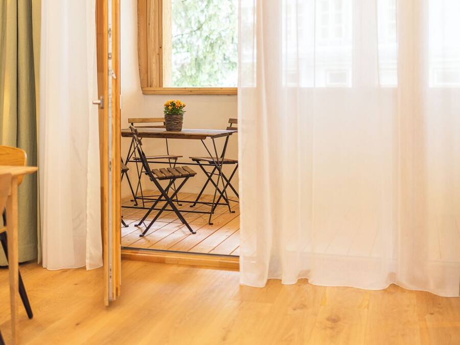 Een open deur die leidt naar een eetgedeelte met een houten tafel en stoelen, een raam met uitzicht op groen en een houten plafond.