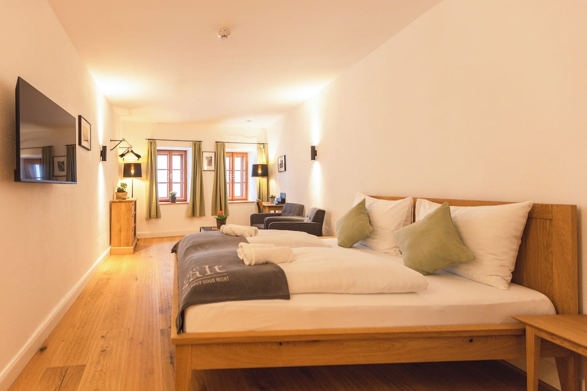 Een slaapkamer met een houten bed, een bureau, een stoel, een aan de muur bevestigde televisie en een raam met gordijnen.