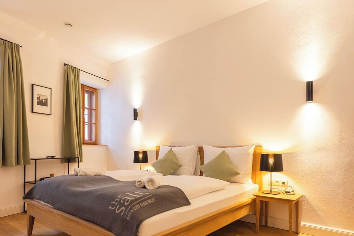 Een slaapkamer met een houten bed, twee nachtkastjes met lampen, een raam met gordijnen en een schilderij aan de muur.