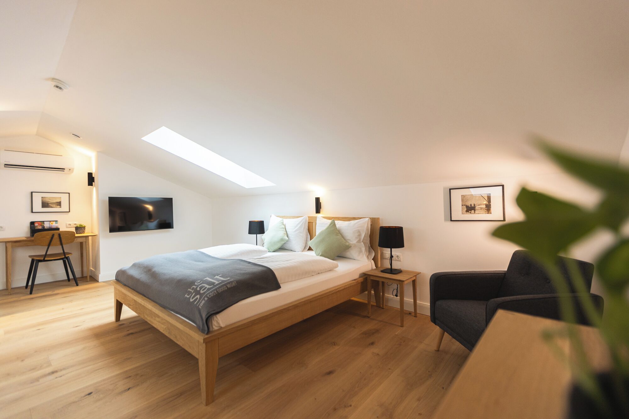 Ein modernes Schlafzimmer mit Oberlicht, einem Bett mit grauer Decke, einem Schreibtisch mit Stuhl und einer Pflanze.