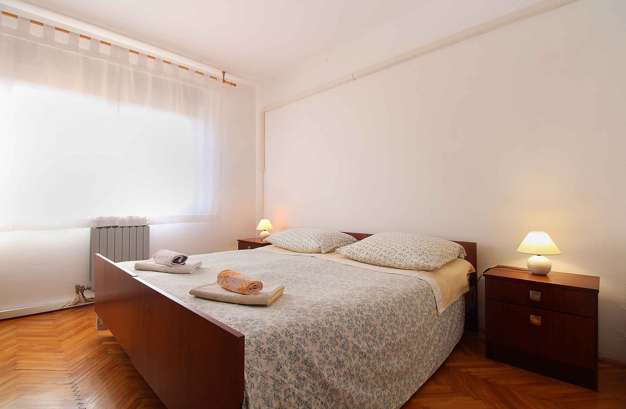 A bedroom with a wooden bed, two bedside tables with lamps, a radiator, and a window with curtains.