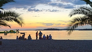 Een strand met mensen die zitten en staan, palmbomen en een zonsondergang boven het water.