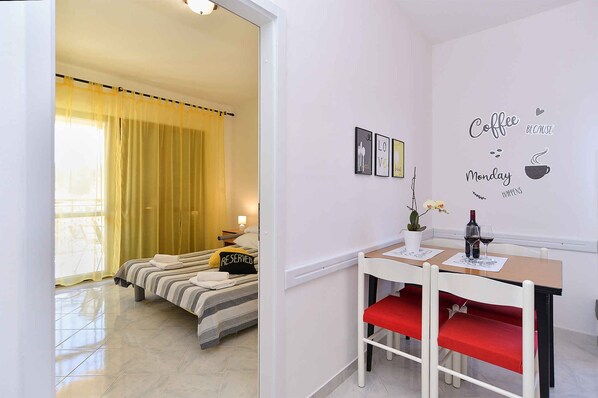 Ett hotellrum med en säng, ett skrivbord och en stol. Det finns ett fönster med gardiner, en vägg med en kaffetematisk dekoration och ett litet bord med en vas och en flaska.