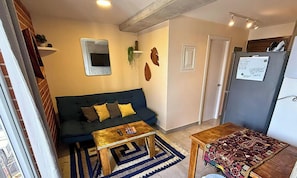 Un salon comprenant un canapé, une table basse, un tapis à motifs, une petite table avec un tapis, et un réfrigérateur sur lequel est accrochée une note.