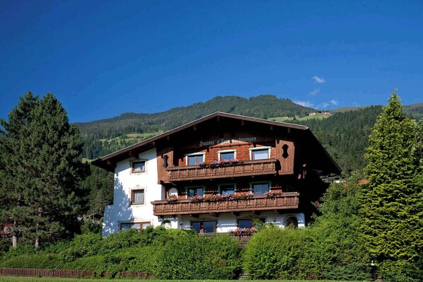 A traditional chalet-style building with multiple balconies, surrounded by lush greenery and mountains in the background.