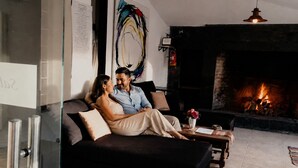 Una pareja en un sofá de color oscuro en una sala de estar con chimenea, una pared de vidrio y una lámpara colgante moderna.